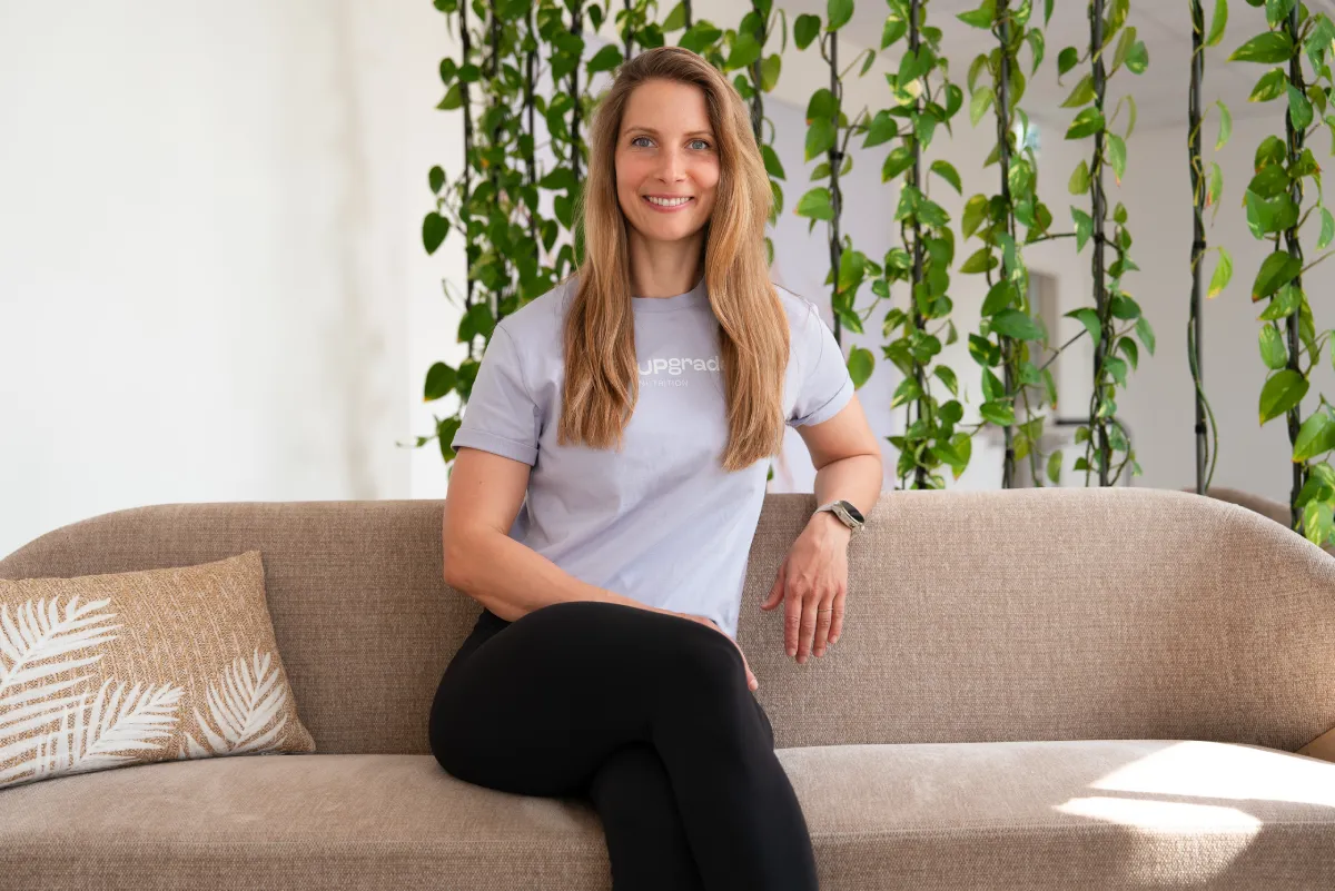 Portrait von Carina Kohlhauser, sitzend auf einer beigen Bank, Pflanzen im Hintergrund